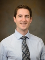 Professional headshot of a man wearing a light blue dress shirt and a plaid tie.