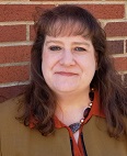A woman with long brown hair and a friendly smile, wearing an orange blouse and a brown blazer, stands against a brick wall.