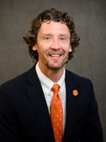 Professional headshot of a man with curly hair, wearing a navy suit and an orange tie, featuring a Clemson University logo on his lapel.