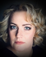 A close-up portrait of a woman with blonde, curly hair and striking blue eyes, exhibiting a natural makeup look.