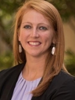 Professional headshot of a woman with red hair and earrings, smiling in a outdoor setting.