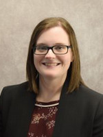 Professional headshot of a woman with shoulder-length brown hair, wearing glasses and a black blazer over a maroon blouse, smiling against a neutral background.