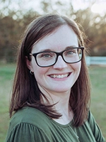 A smiling woman with glasses, standing outdoors in a natural setting, wearing a green shirt.