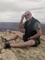 A man sitting on a rocky ledge at a canyon, wearing shorts and a vest, with a contemplative expression.