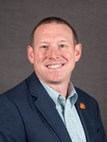 Professional headshot of a man in a suit, smiling against a neutral gray background.