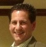 A smiling man with dark spiky hair dressed in a suit and checkered shirt.