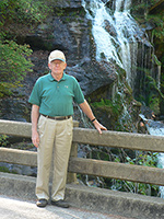 An elderly man in a green shirt and beige pants stands beside a stone railing with a waterfall in the background.