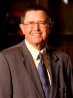Formal portrait of a smiling man in a suit and tie, standing against a bookshelf background.