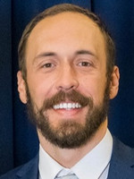 Headshot of a smiling man with a beard, wearing a suit and tie against a blue backdrop.
