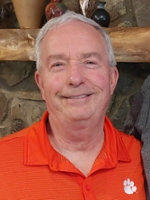 Smiling older man wearing an orange shirt with a Clemson University logo, posing in front of a rustic stone wall.