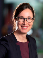 A professional headshot of a woman with glasses and long brown hair, smiling while wearing a dark blazer over a burgundy top.