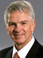 Professional headshot of a man with gray hair, wearing a dark suit and orange tie.