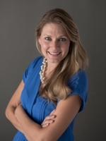 Professional headshot of a woman with long blonde hair wearing a blue blouse and pearl necklace, smiling and standing with her arms crossed.