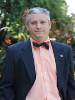 A man in a suit and bow tie stands in a garden with colorful flowers in the background.