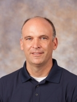 Headshot of a man with short hair, wearing a navy blue polo shirt, smiling against a neutral background.