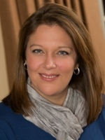 Professional headshot of a woman with shoulder-length brown hair, wearing a blue top and light gray scarf, smiling warmly at the camera.