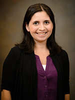 Headshot of a smiling woman with dark hair wearing a purple shirt and black cardigan against a neutral background.