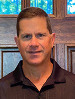 Headshot of a man smiling, wearing a black polo shirt, with wooden doors and greenery in the background.