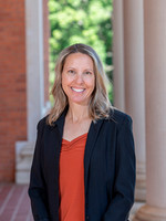 A smiling woman wearing a black blazer and orange top stands in a bright outdoor setting with columns in the background.