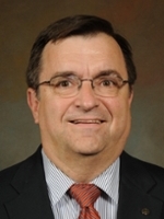 Headshot of a middle-aged man wearing glasses, a dark suit, and a striped tie, smiling at the camera.