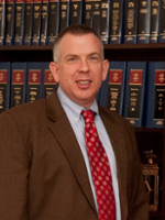 A professional portrait of a man in a brown suit and red tie, smiling in front of a bookshelf filled with legal books.