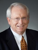 Professional headshot of a smiling older man wearing glasses, a suit jacket, and a patterned tie.
