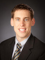 Professional headshot of a smiling man in a suit and tie against a dark background.