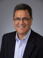 A smiling man in a professional suit and glasses, positioned against a neutral gray background.