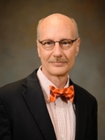 Professional headshot of a man with a mustache and orange bow tie, wearing glasses and a black suit jacket.