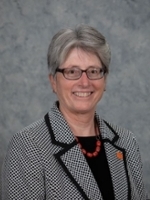 Professional portrait of a woman with short gray hair, wearing glasses and a patterned blazer, smiling against a gray background.