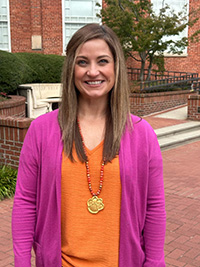 A woman smiling in a pink cardigan over an orange top, standing outdoors with a brick building and greenery in the background.