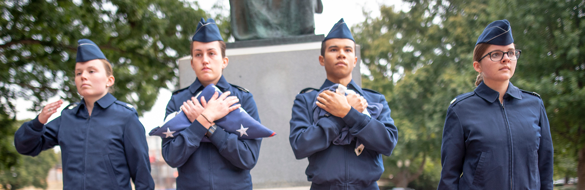 Four cadets in dress uniform standing at attention and holding folded US flag in front of Tillman Hall.