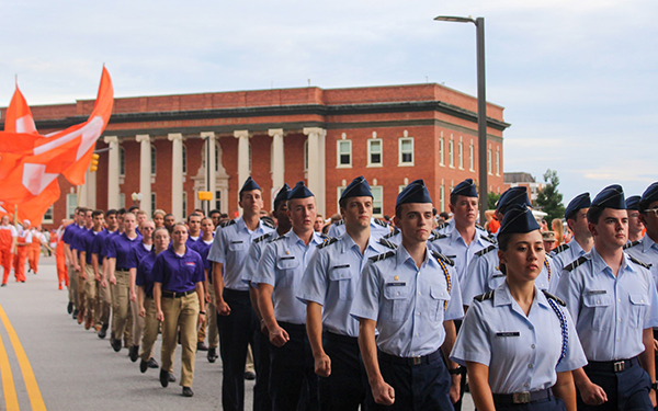 Members of a military cadet corps marching in a parade, wearing uniforms and showcasing school spirit with orange flags.