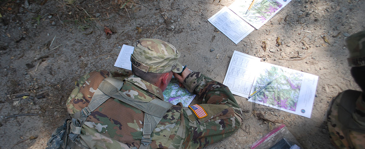 Cadets in camouflage laying on ground drawing maps.