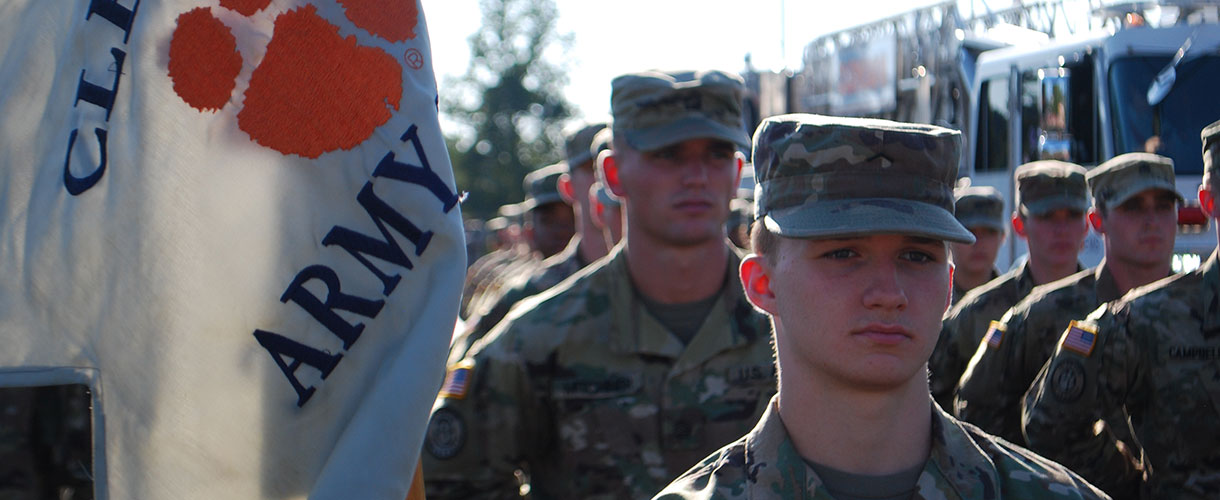 Cadets in camouflage marching in parade with Clemson flag.