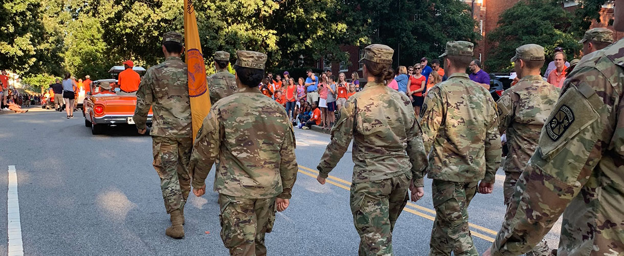 Cadets in camouflage marching in parade with Clemson flag.