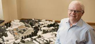A smiling architect stands beside a detailed architectural model of a campus layout.