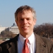 Portrait of a man in a suit with a tie, smiling outdoors with a blurred cityscape in the background.