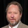 Professional headshot of a man with a beard, wearing a suit and tie, smiling at the camera.