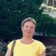 A woman in a yellow dress stands in front of greenery and a building.