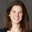 Professional headshot of a smiling woman with long brown hair against a grey backdrop.