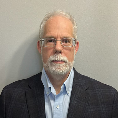 A professional headshot of a man with gray hair and a beard, wearing glasses and a dark blazer over a light blue shirt.