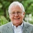 Smiling man wearing glasses and a light gray checkered blazer, standing outdoors amidst greenery.