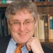Professional headshot of a smiling man with gray hair, wearing glasses and a blue shirt with an orange tie, against a background of bookshelves.