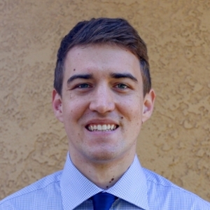 A smiling young man wearing a collared shirt and tie in front of a textured yellow wall.