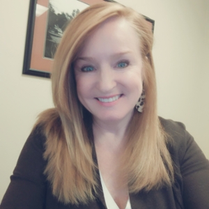 Professional portrait of a smiling woman with long, straight hair, wearing a dark blazer and earrings.