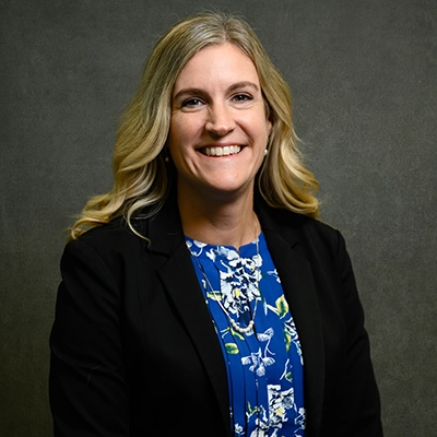 Profile photo of a woman with long blonde hair, wearing a black blazer and a blue floral dress, smiling in front of a gray background.