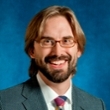 Professional headshot of a man with glasses and a beard, wearing a suit and tie, smiling against a blue background.