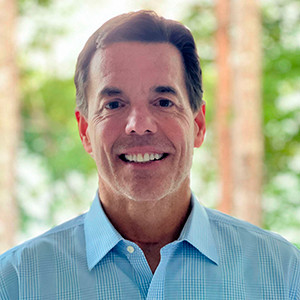 Headshot of a man in a light blue shirt smiling, with a blurred green background.