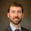 Headshot of a man with a beard, wearing a suit and tie, smiling against a neutral background.